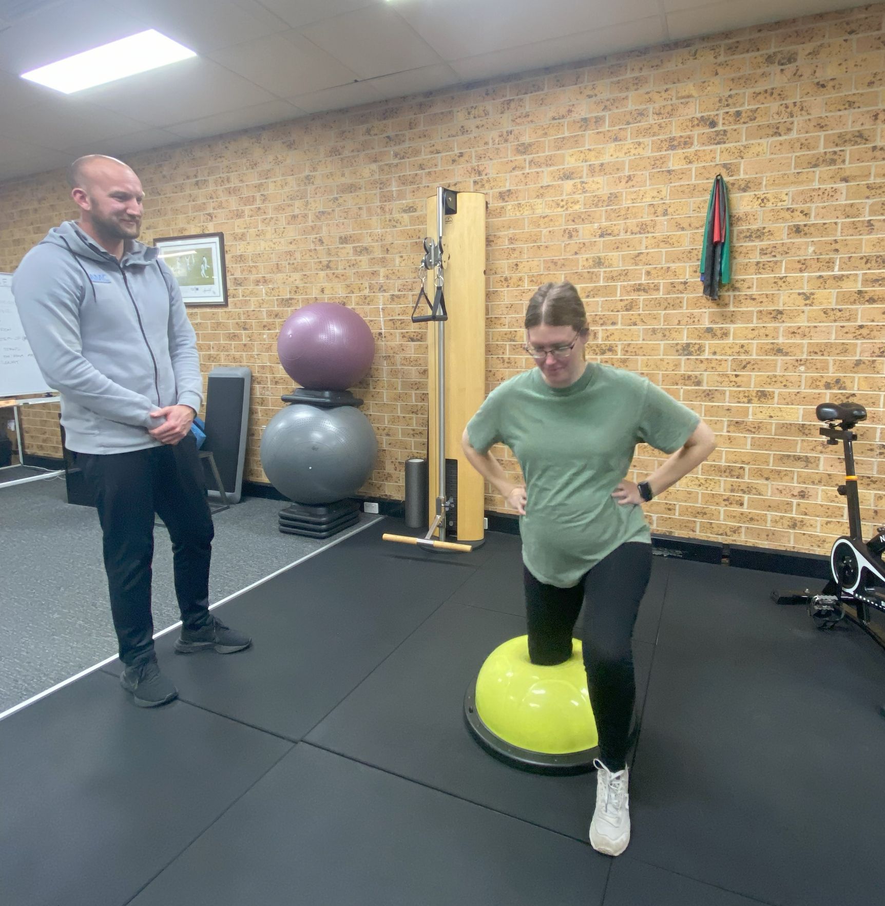 A woman is standing on a balance ball in a gym while a man watches — AMC Exercise Physiology in Maitland, NSW