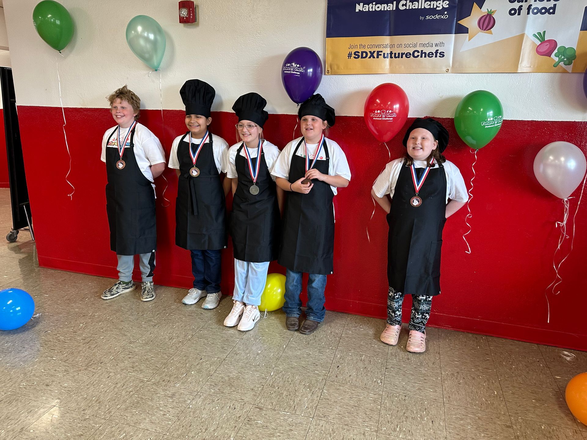 A group of children wearing aprons and chef hats are standing in front of balloons.