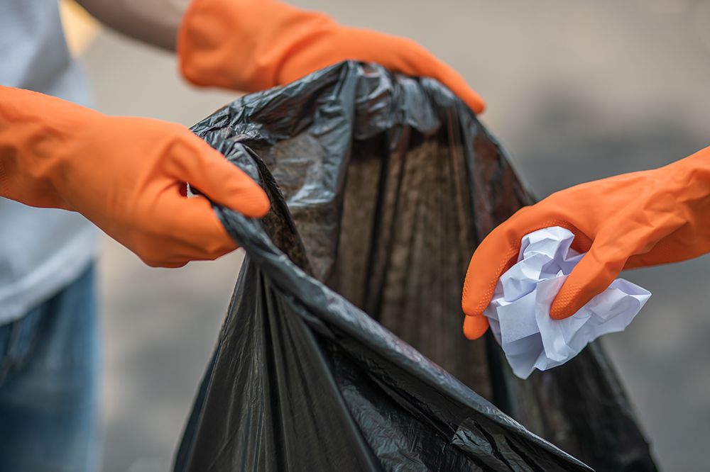 A person wearing orange gloves is putting a piece of paper in a trash bag.