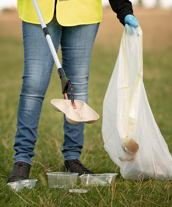 A person is picking up trash in a field with a shovel and a bag.