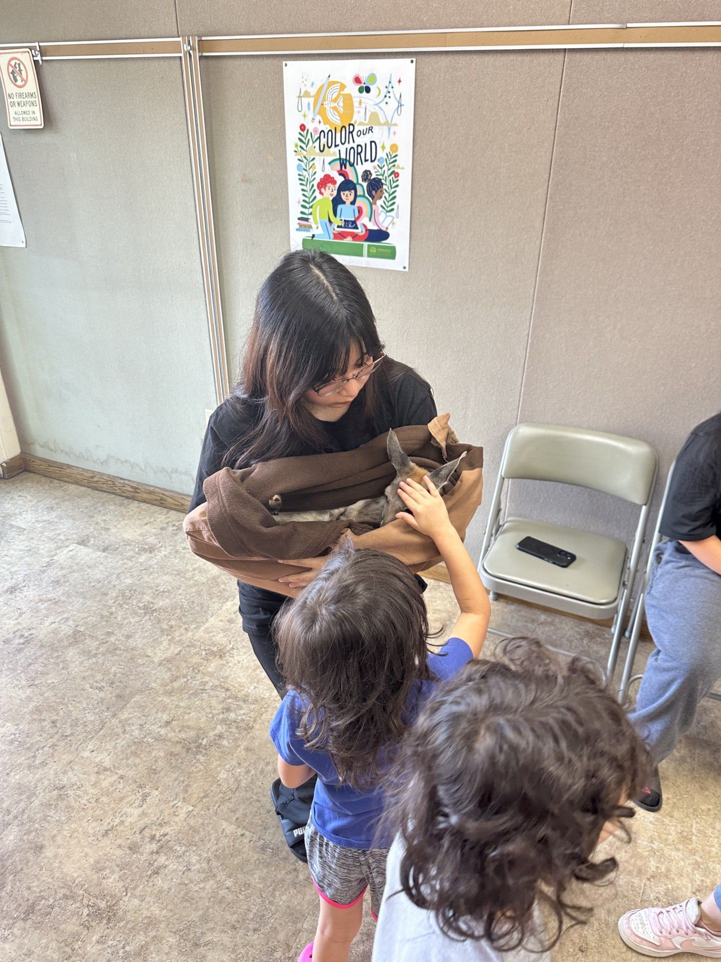 A girl holding a baby kangaroo wrapped in a brown blanket while other kids are petting it