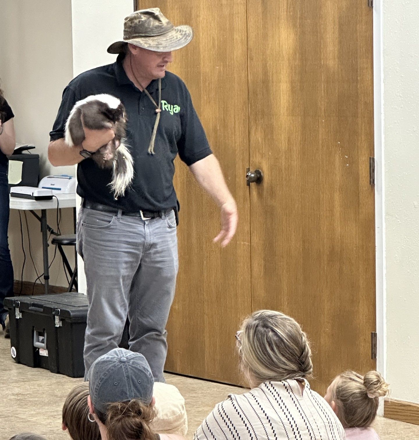 A man in a hat holds a black and white skunk, giving a presentation to children sitting on the floor.