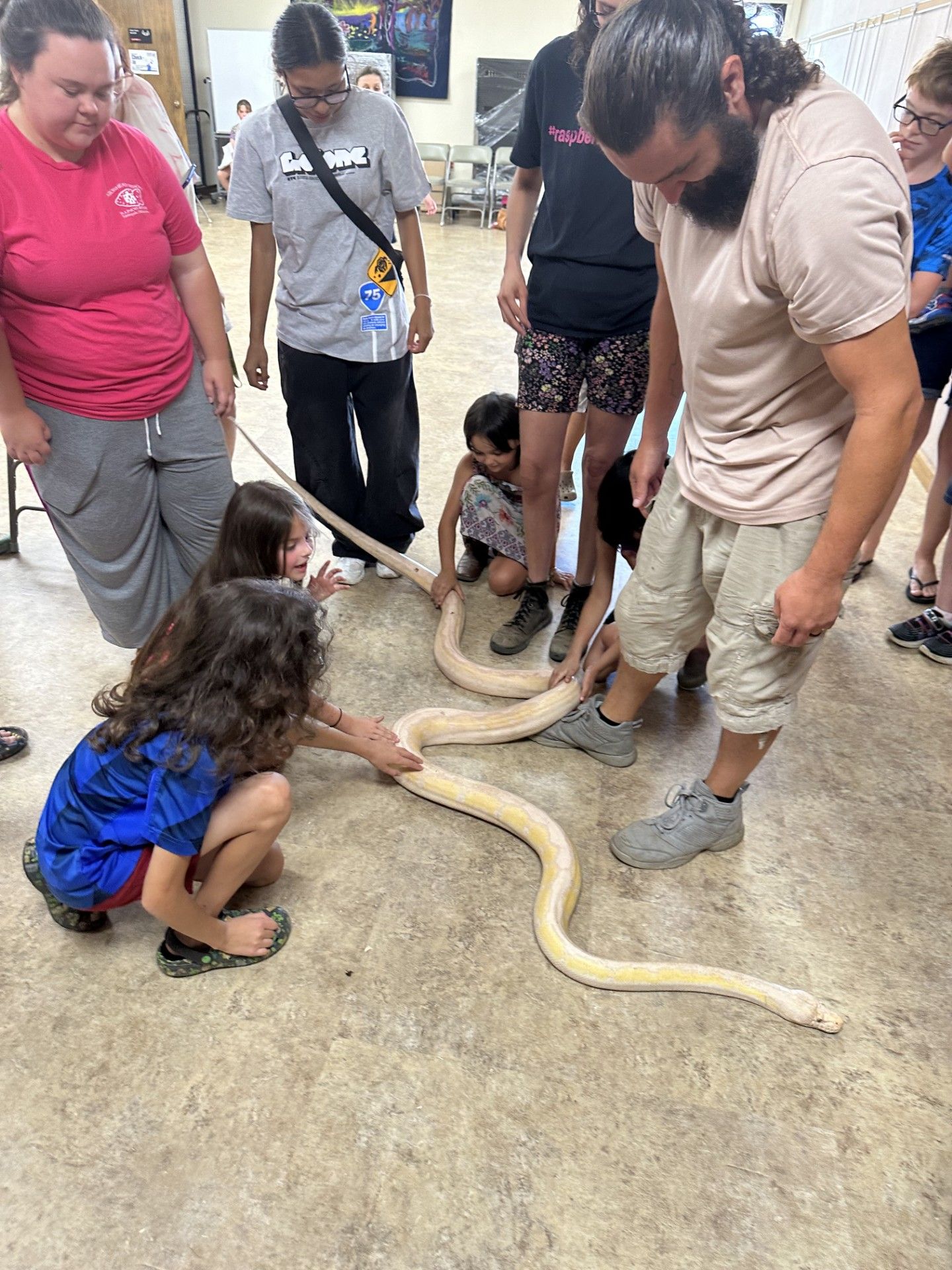 People, including children, gather around a large, pale snake on a floor indoors, likely at an educational event. They're touching it curiously.