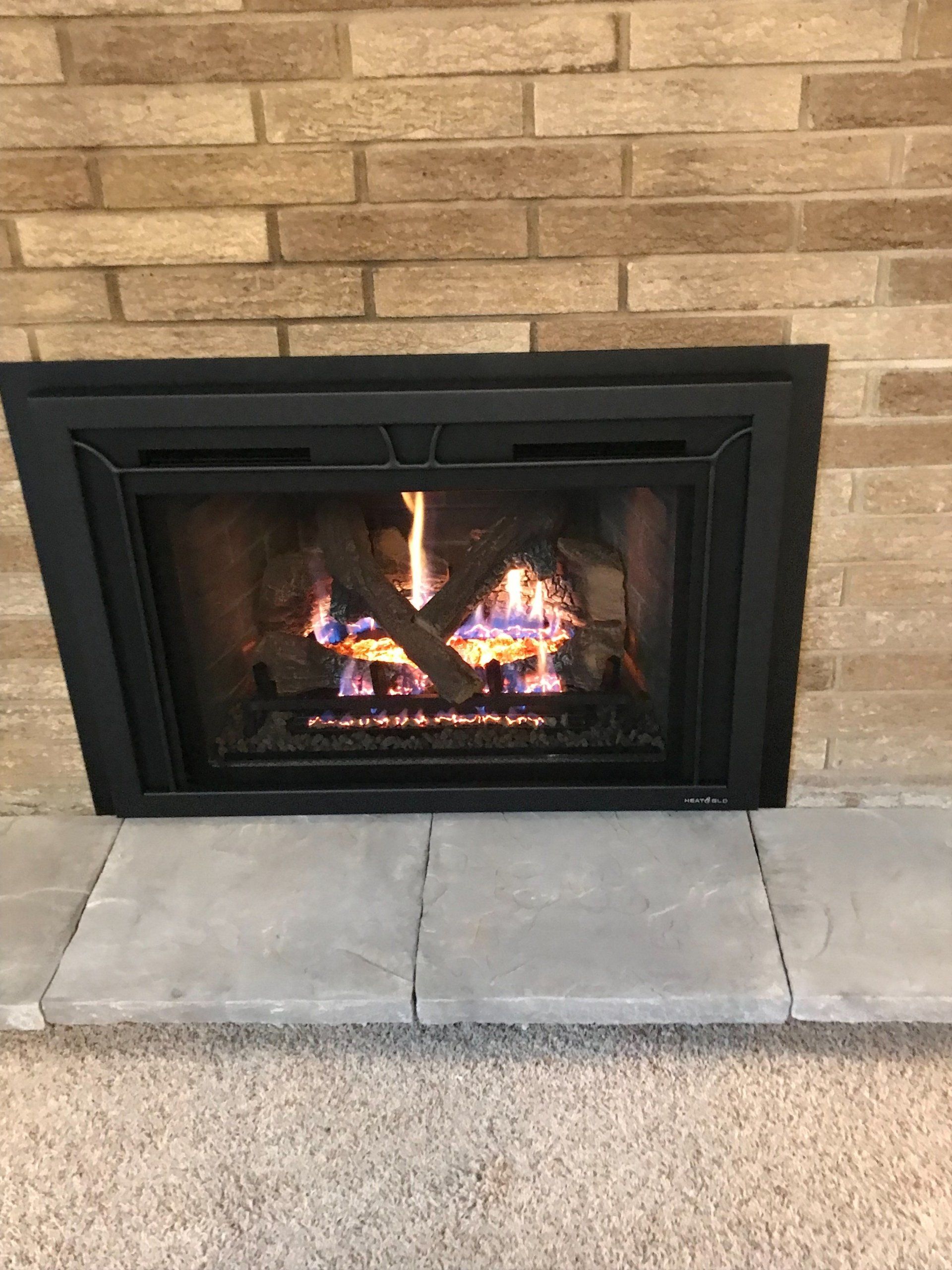 A gas fireplace with flames burning inside, framed by a black border, set against a brick wall.