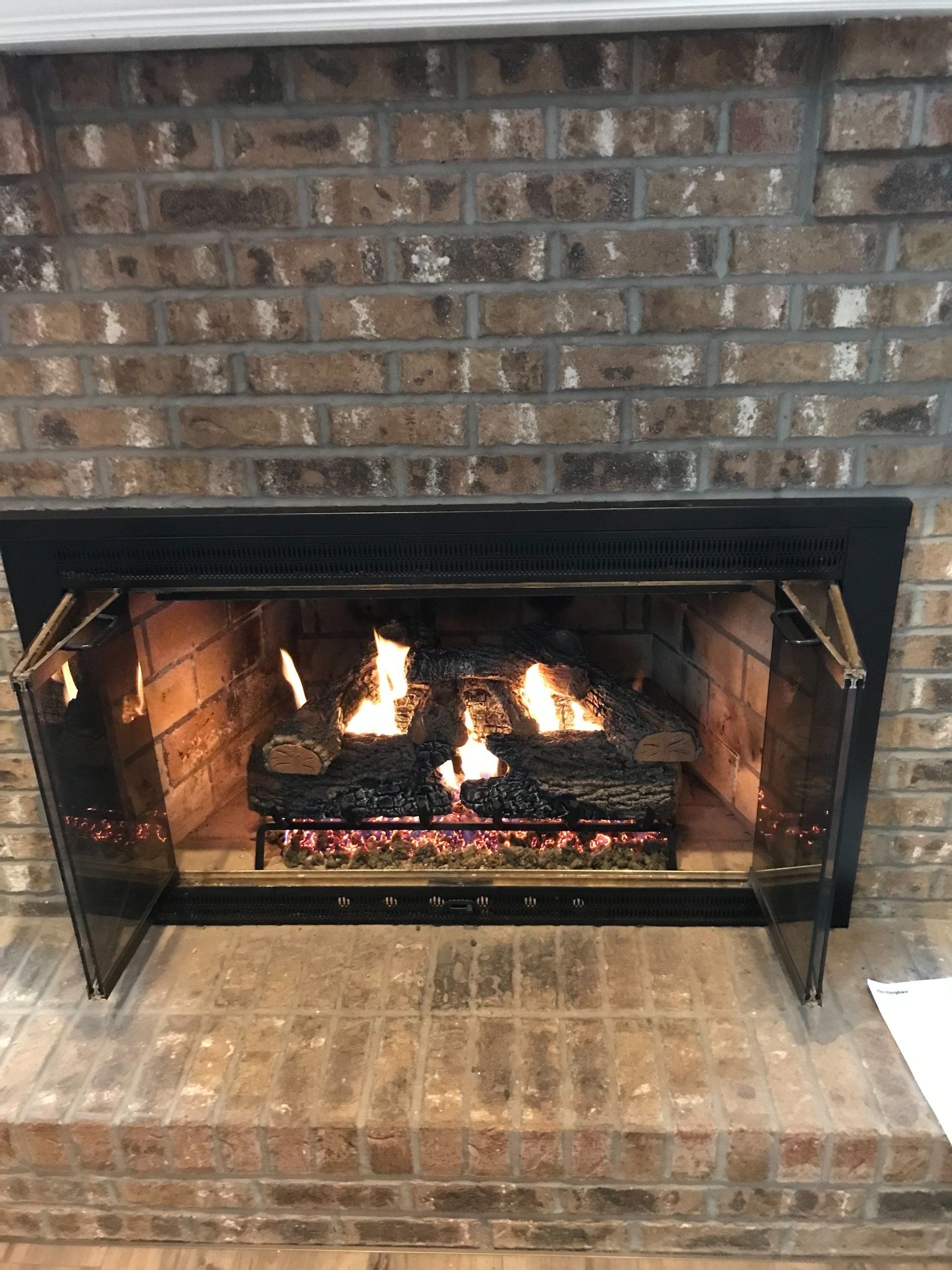 Fireplace with burning logs, glass doors open, set in brick, on a brick hearth.