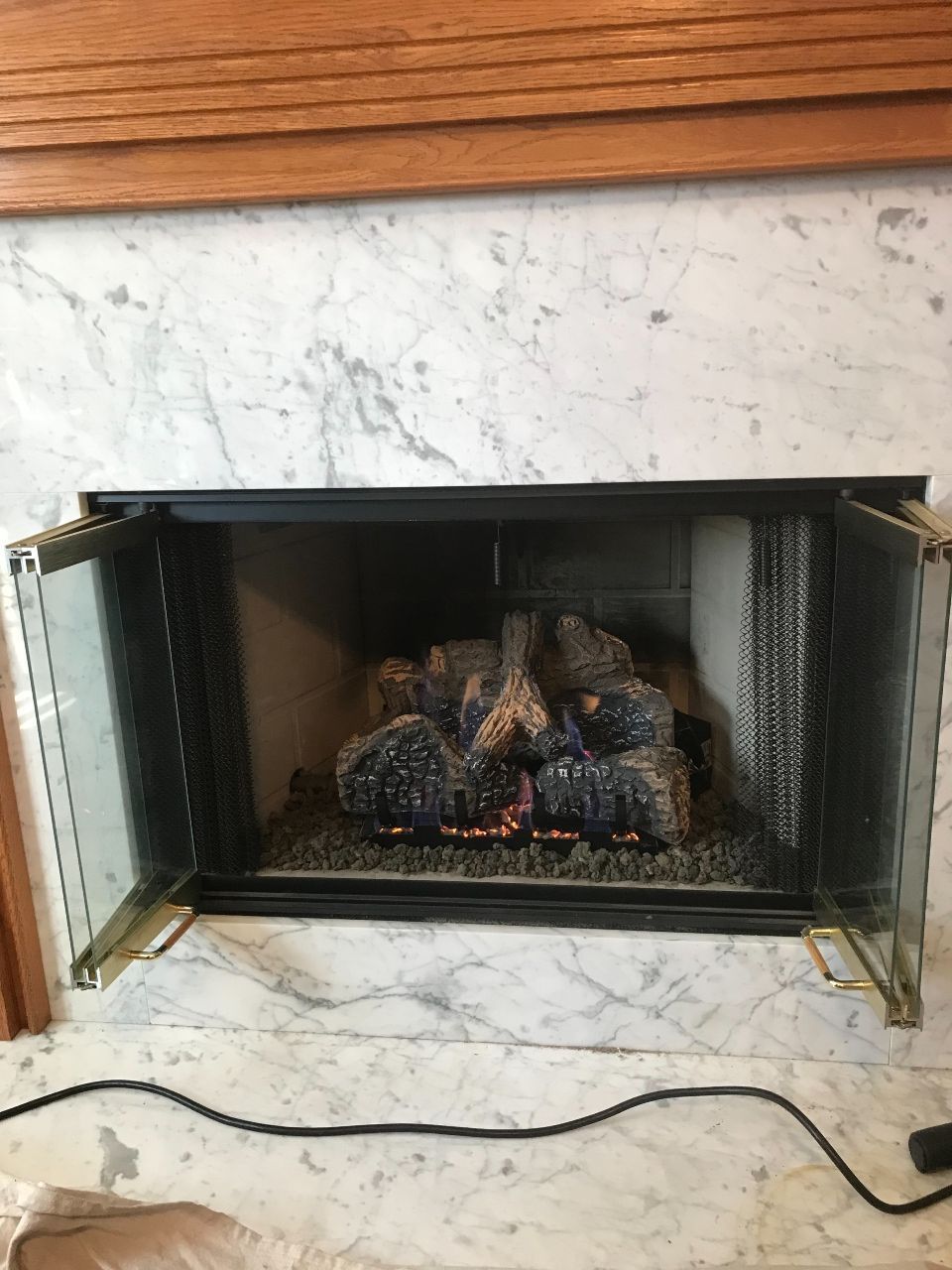 Fireplace with glass doors, lit gas logs, set in marble and framed by wood.