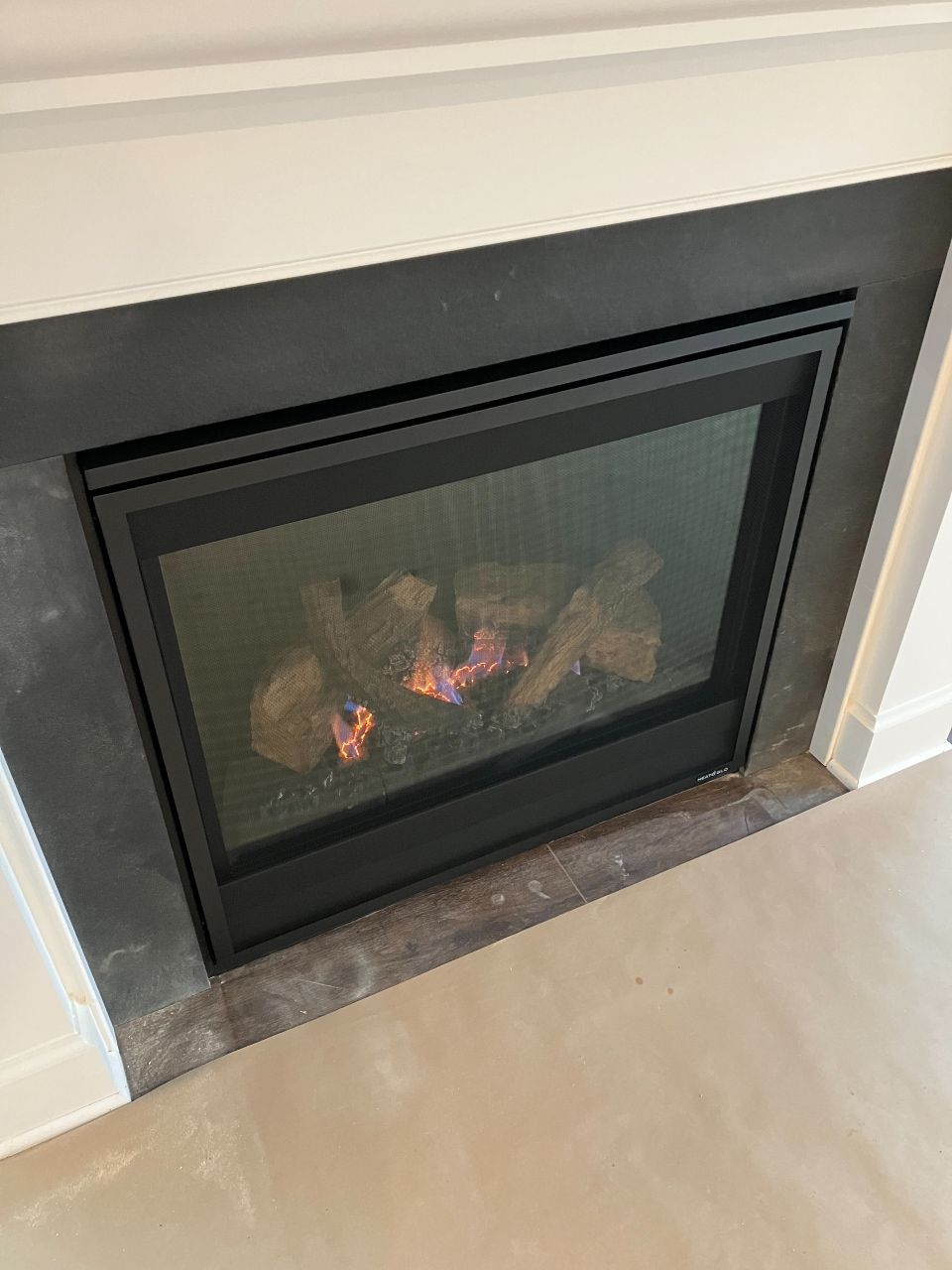 Fireplace with black frame, lit logs, surrounded by dark stone and white trim.