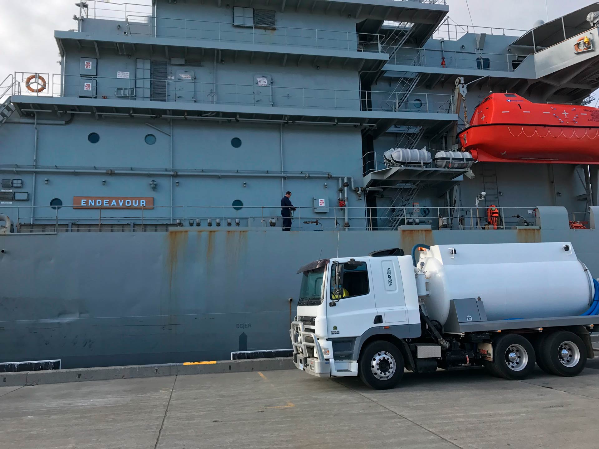 A white tanker truck fueling a large gray naval ship at a dock.
