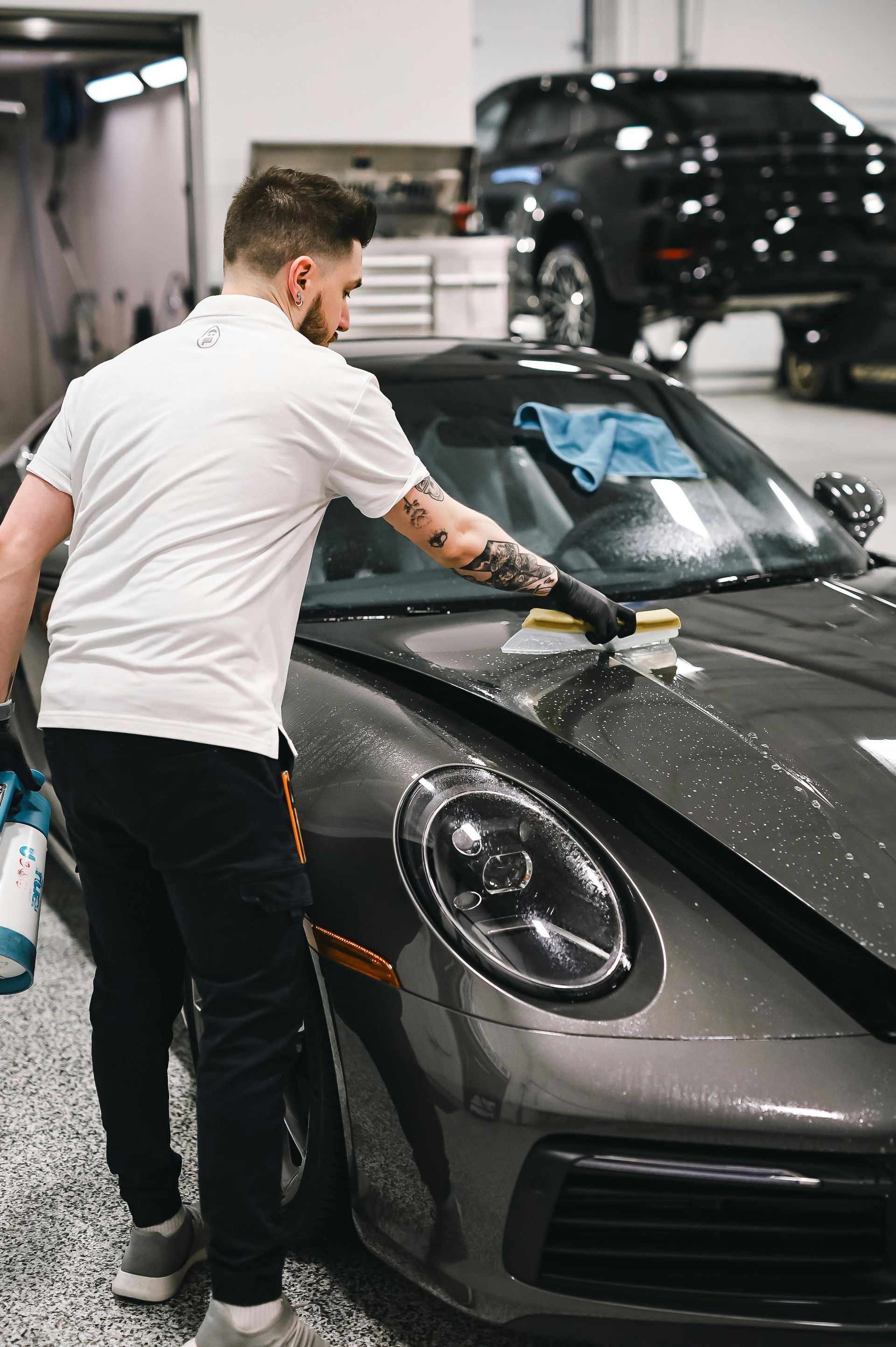 Man polishing a black sports car in a garage, with another vehicle in the background