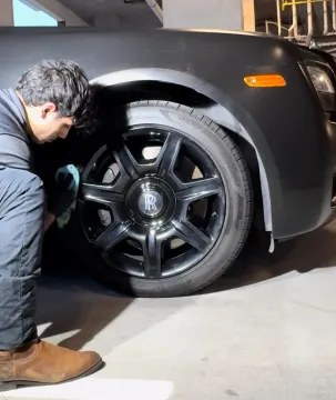 Mechanic crouching beside a black car, inspecting the front wheel in a garage.