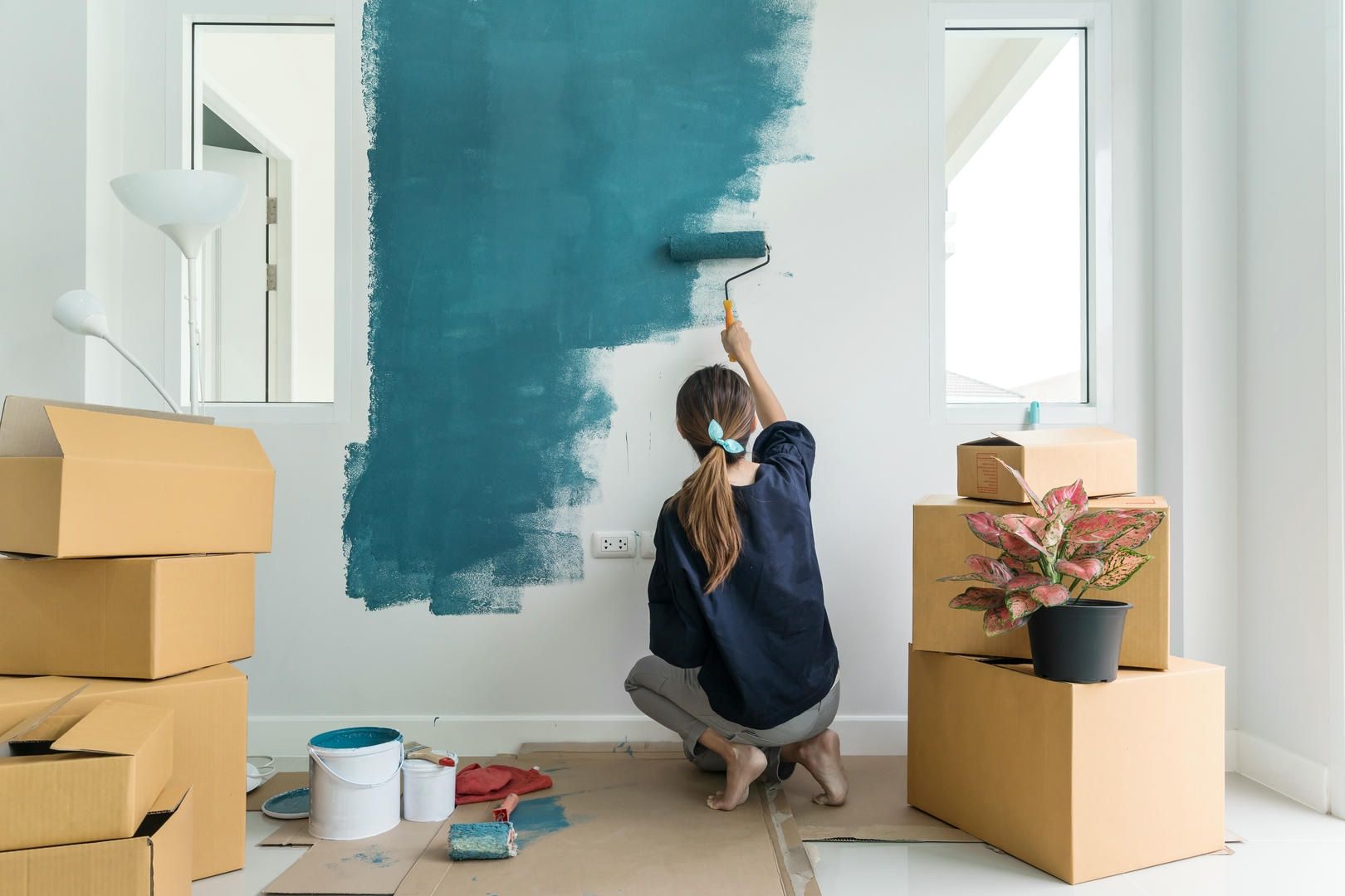 A woman is kneeling on the floor painting a wall with a paint roller.