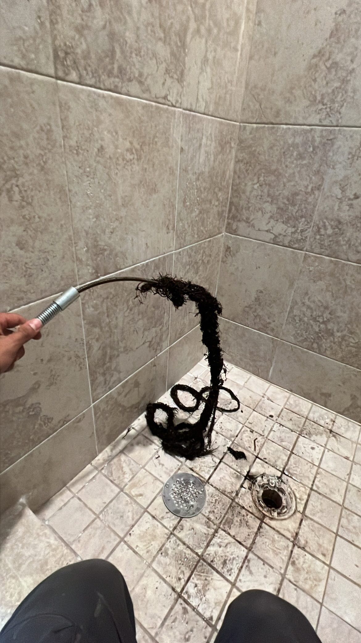 A drain snake pulls out a large clump of dark hair from a shower drain in a tile-walled shower.