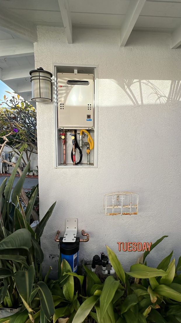 Outdoor water heater installation with pipes, filter, and plumbing against a stucco wall.