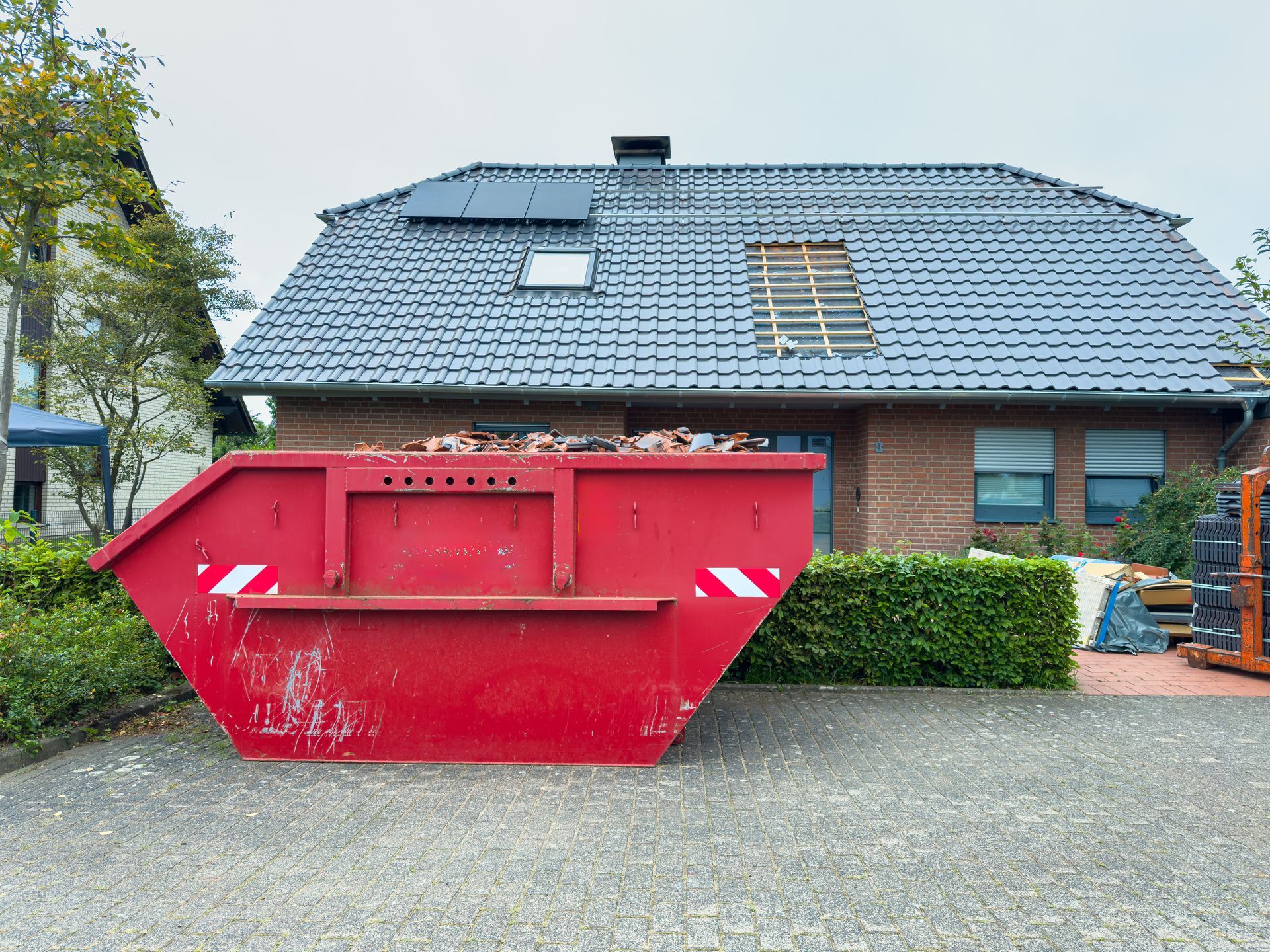 A bright red roll-off residential dumpster stands out against a suburban house with gray shingles.