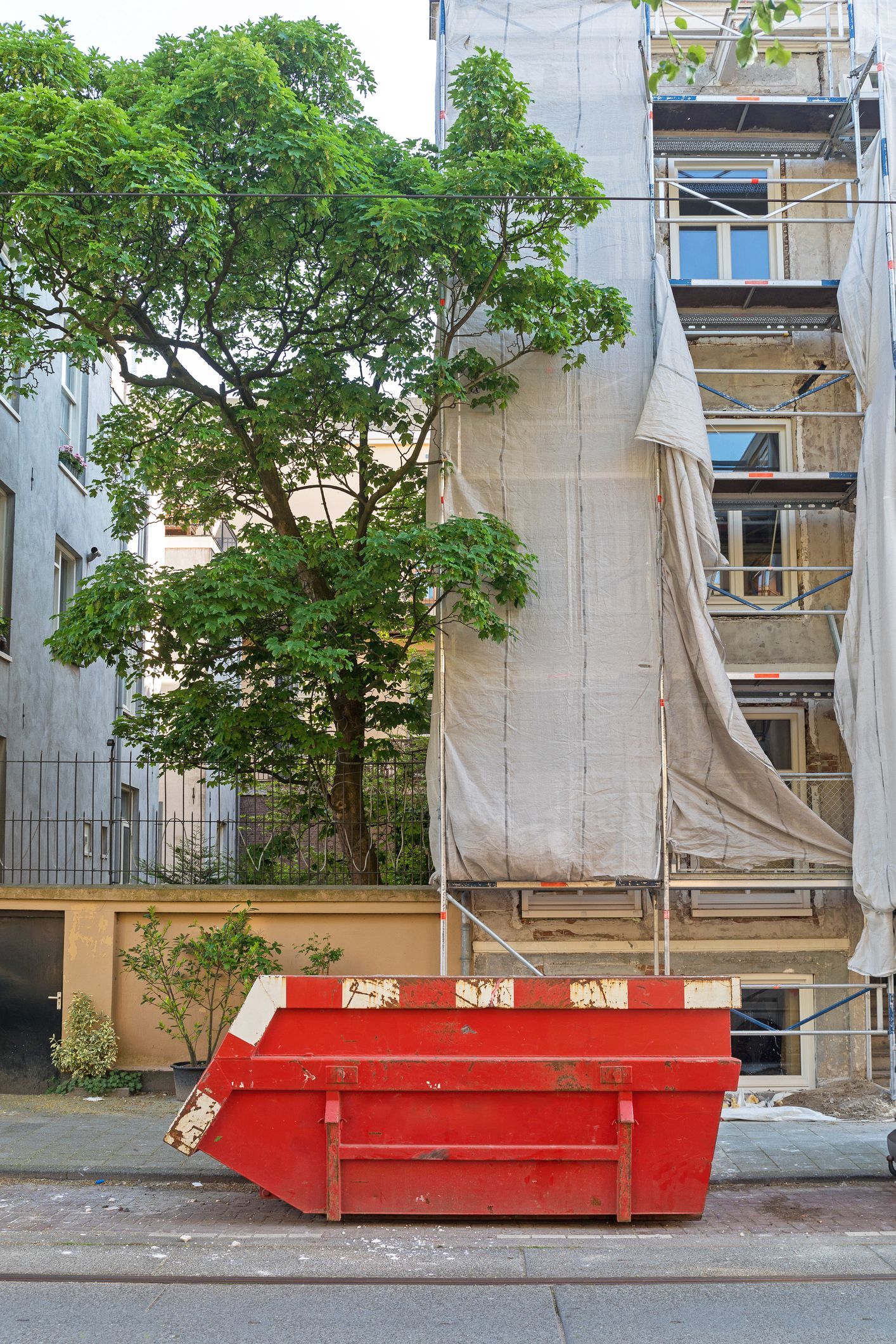 Red dumpster on a sidewalk in front of a building with scaffolding and a large tree.
