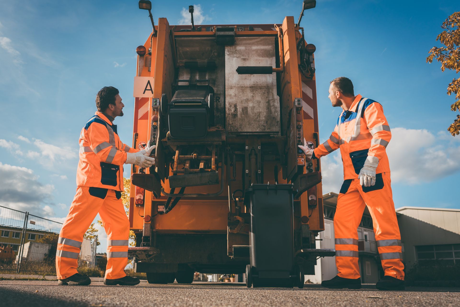 Two sanitation workers in orange uniforms operating a garbage truck outdoors.