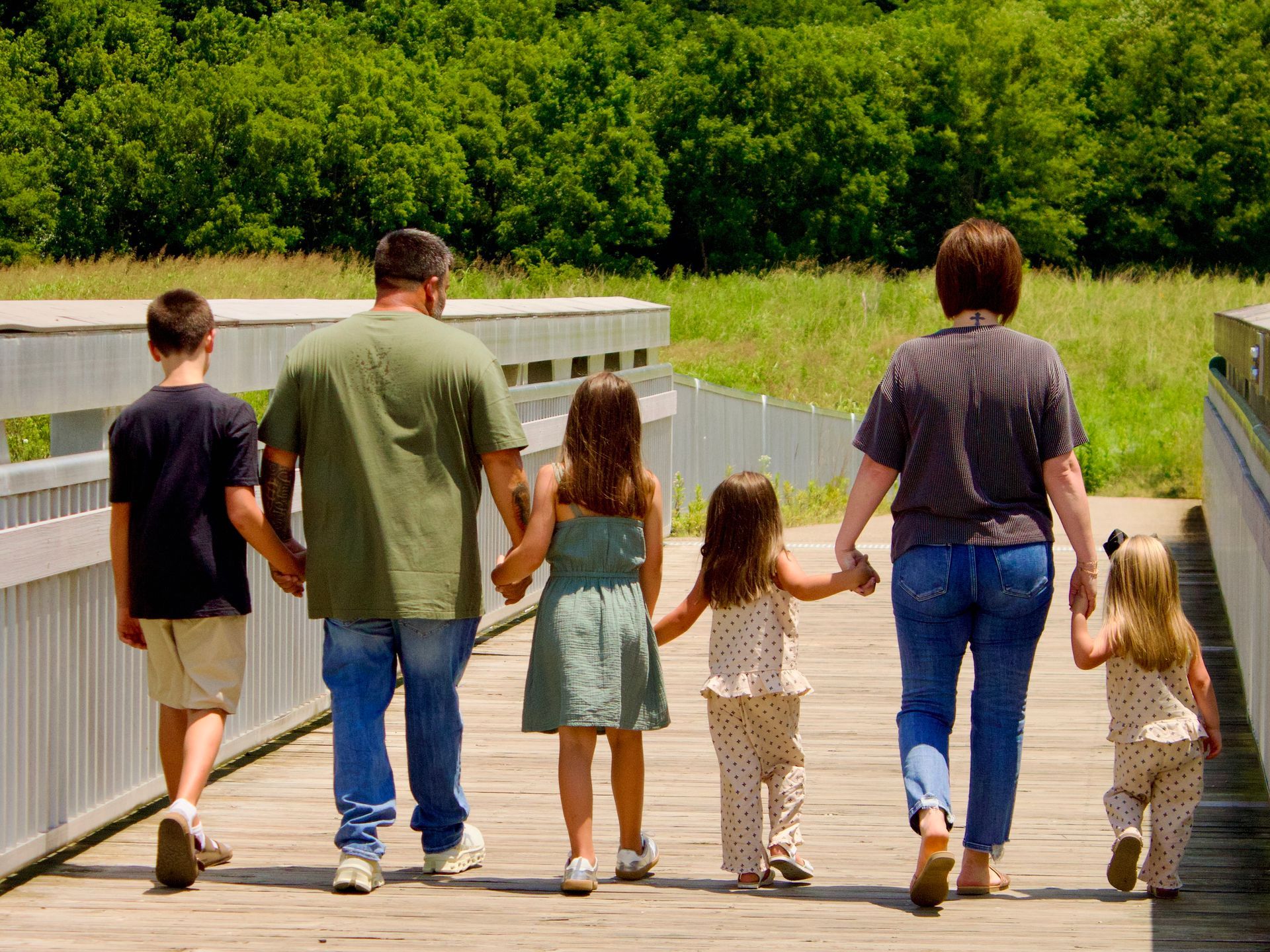 Family of six walking hand-in-hand on a wooden bridge in a park, surrounded by greenery.