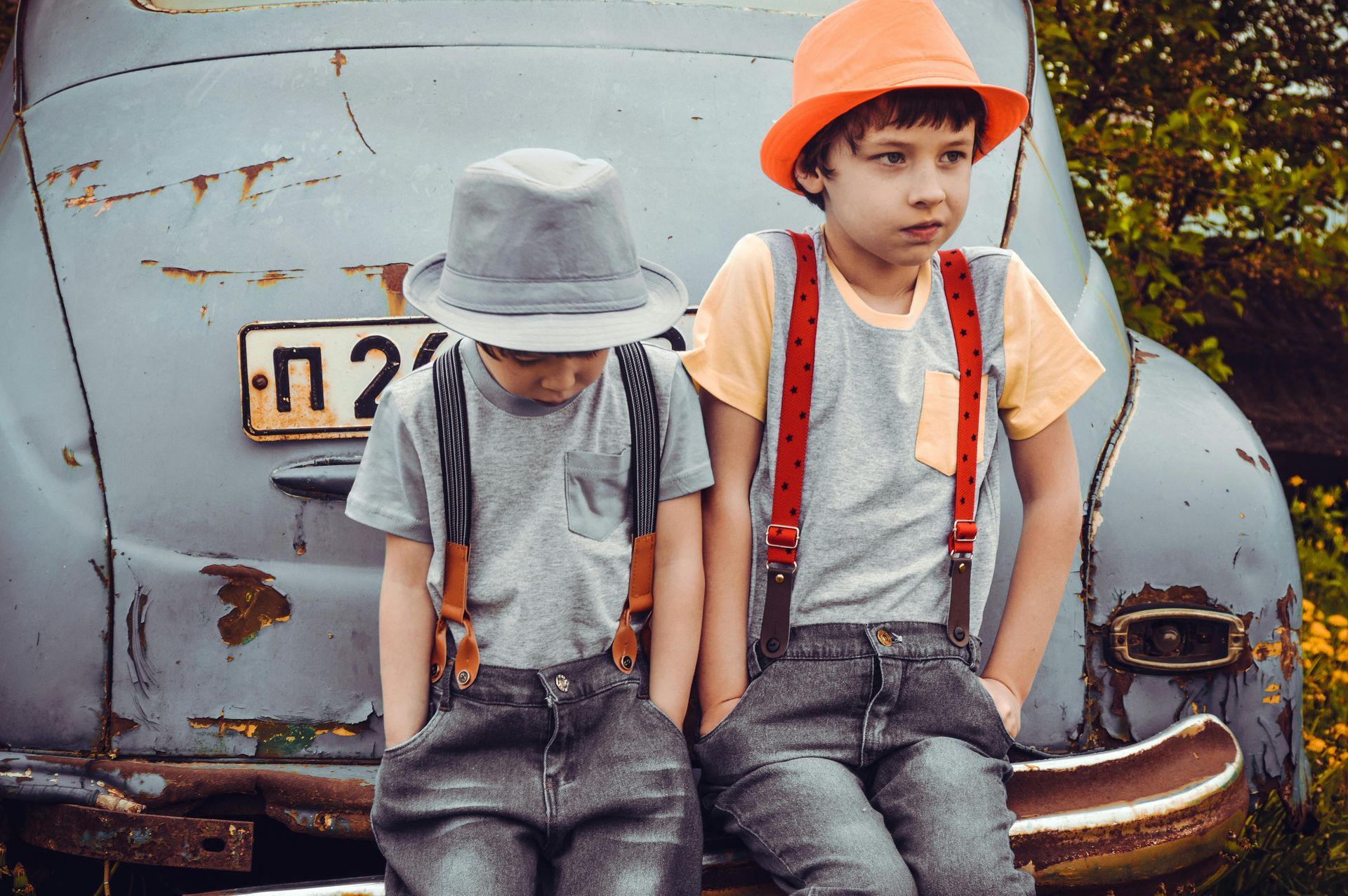 Two boys in suspenders and hats sitting on an old car, looking down.