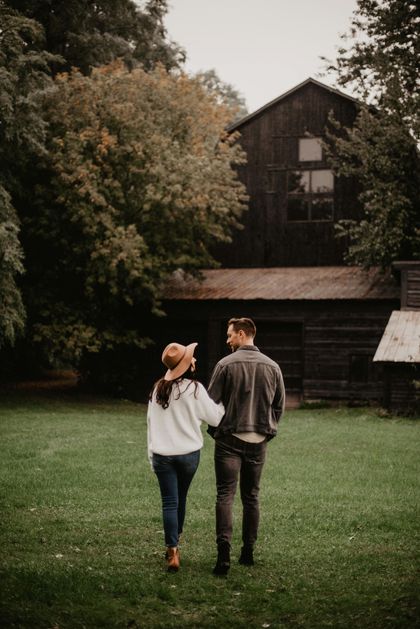 Couple walking in front of a barn. Woman in hat, man in denim jacket; green grass, fall trees.