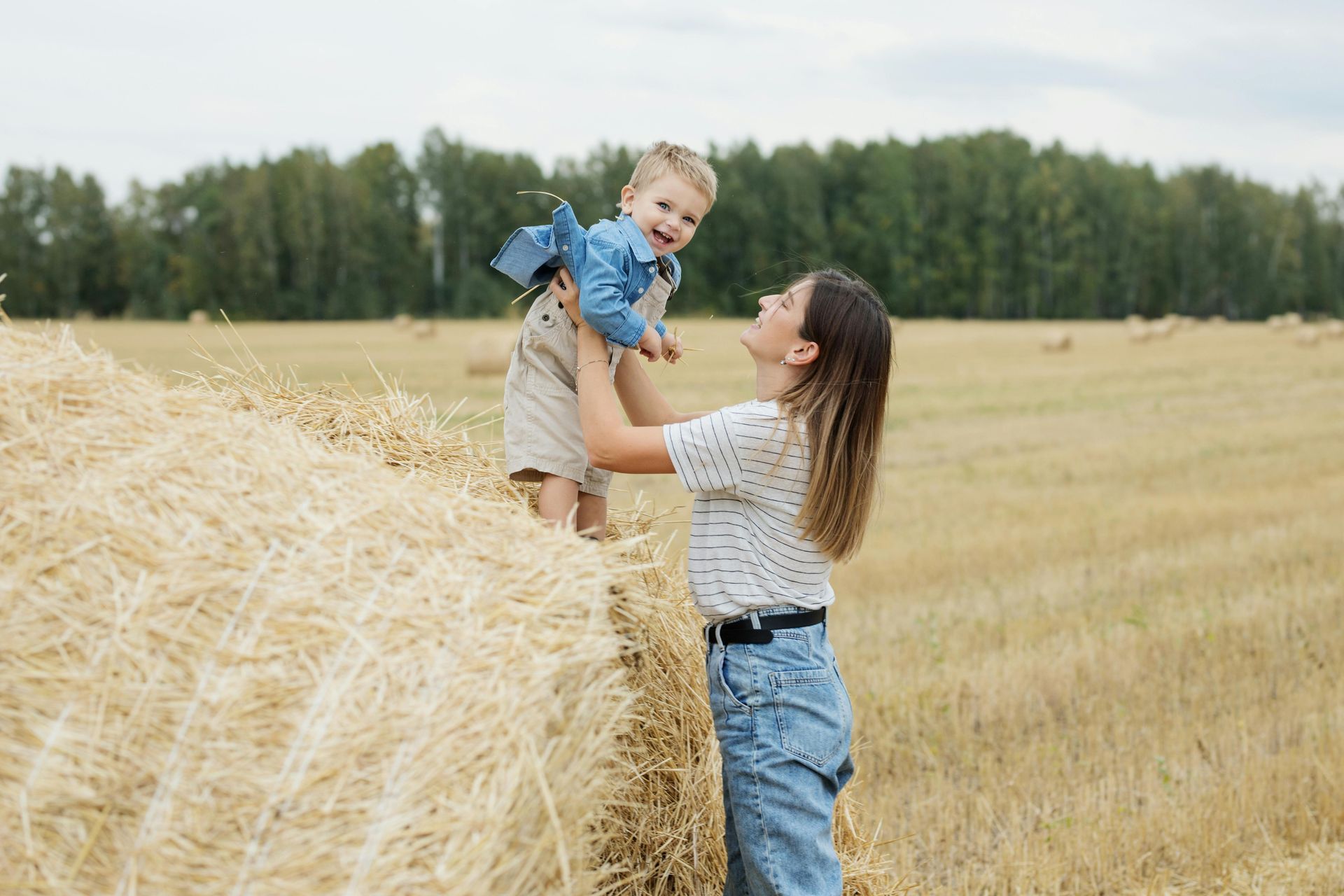 Mother lifting a child in a field of hay bales. They are smiling; the setting is a sunny day.