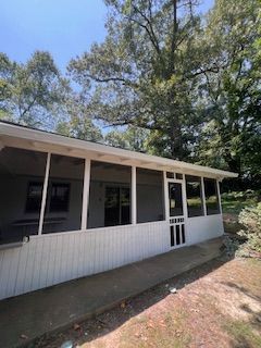 White screened-in porch with windows and door. Concrete walkway. Green trees and blue sky background.