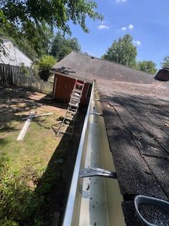 Gutter cleaning on a roof. A ladder leans against the building. The sky is blue, and trees are in the background.