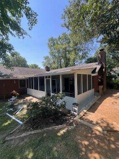 Screened porch attached to a brick house, surrounded by trees. A person stands near the chimney.