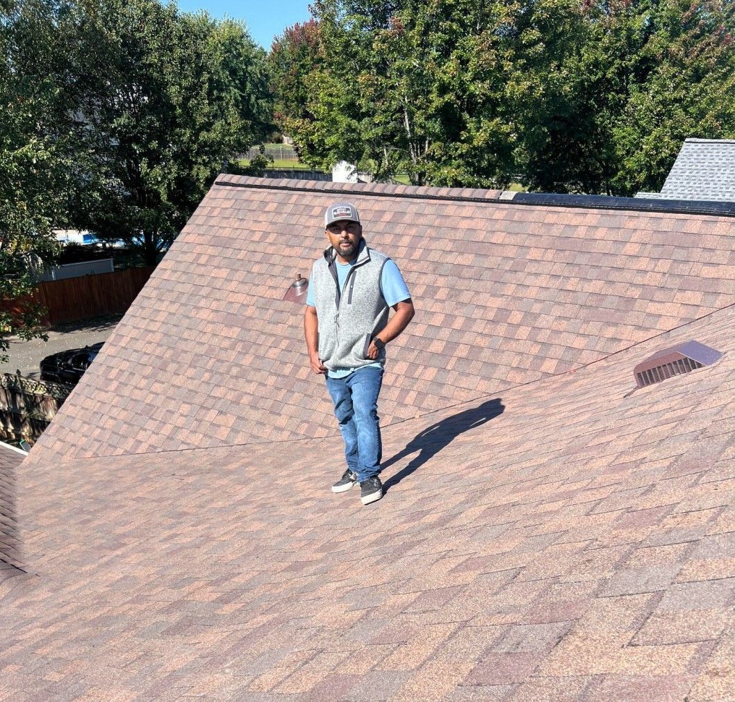 Man stands on a brown shingled roof on a sunny day, wearing a gray vest and jeans.