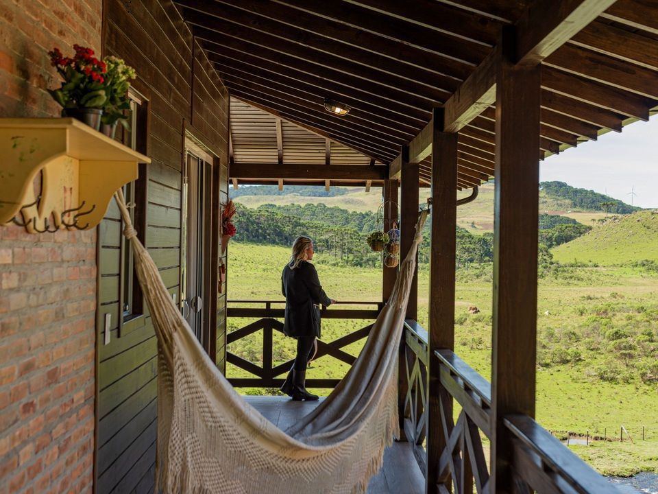 Mulher em pé na varanda de uma cabana, olhando para as colinas verdes. Rede, corrimão de madeira e parede de tijolos.