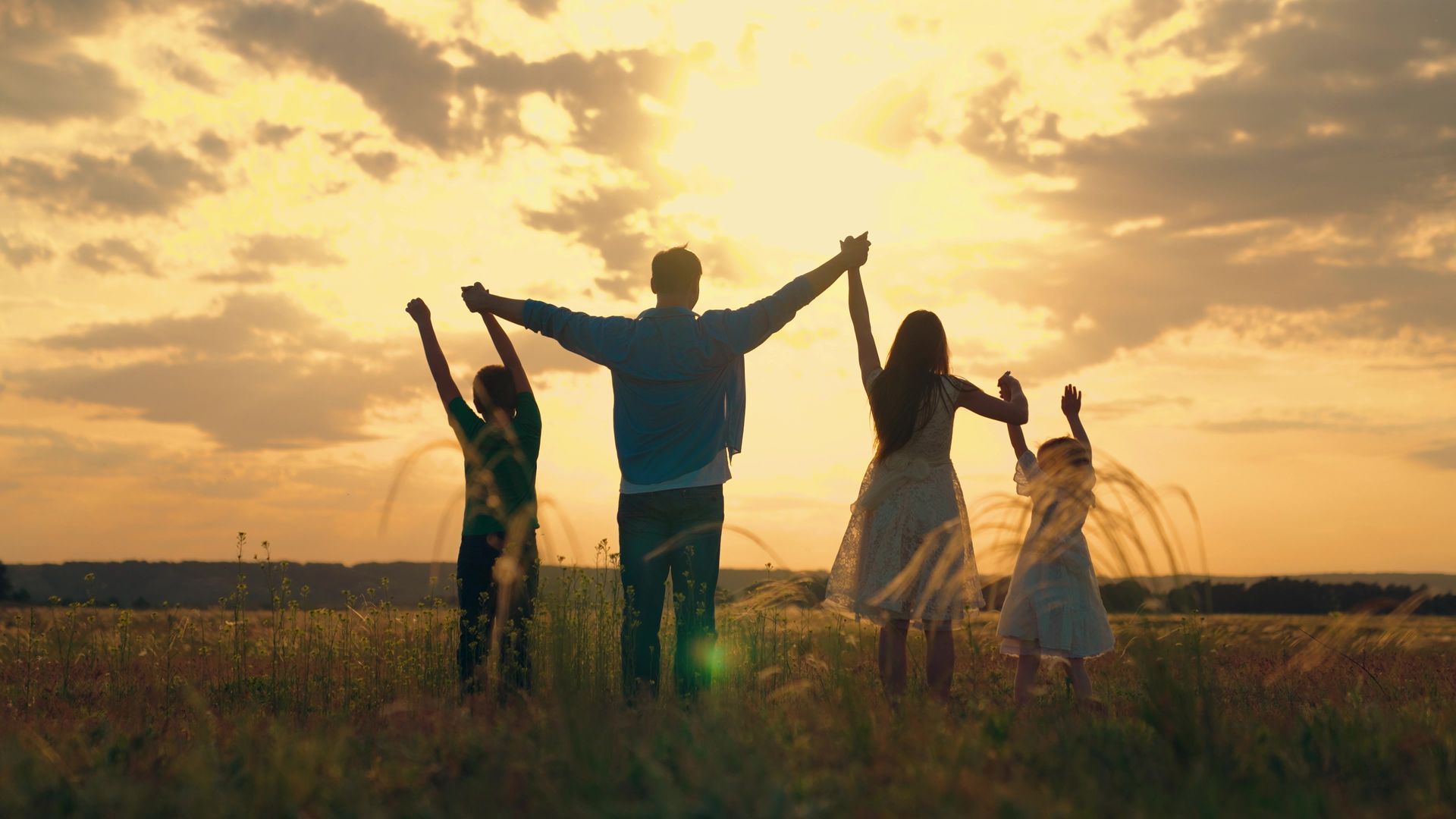 A family silhouette standing in a field at sunset, holding hands with arms raised toward the sky.