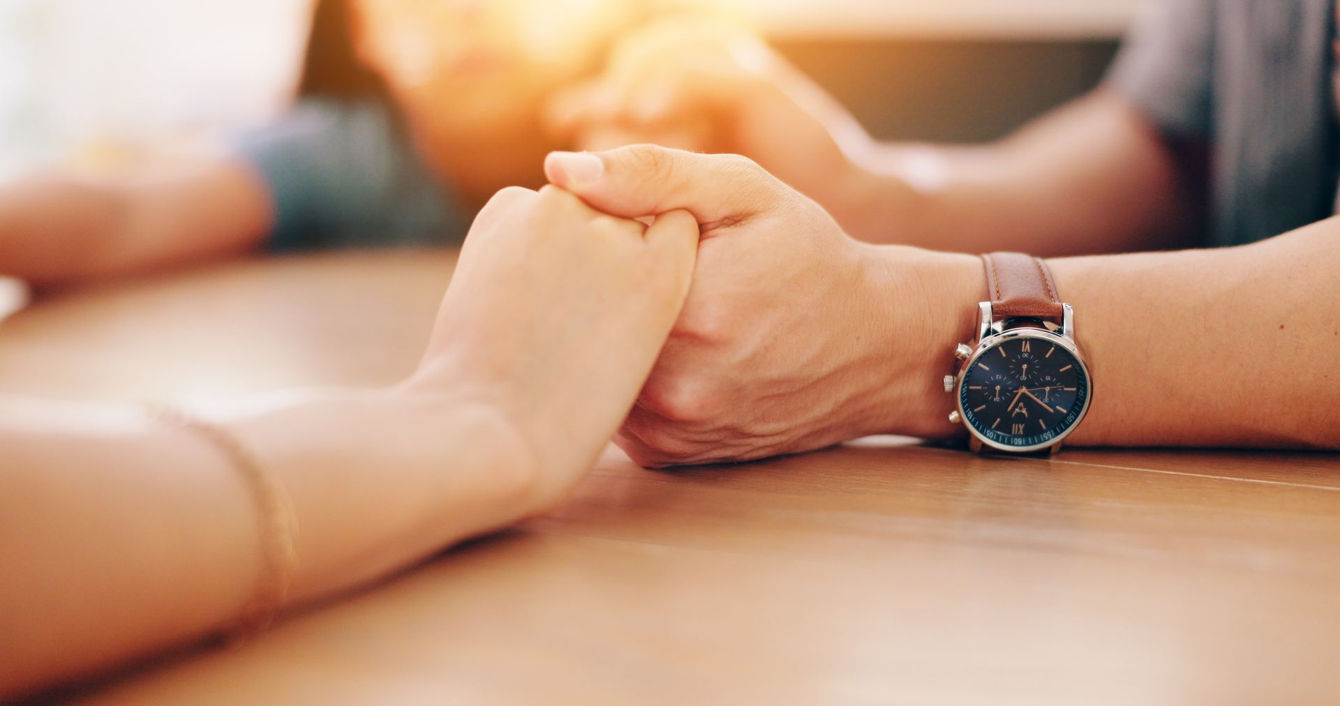 Two people holding hands over a wooden table, one wearing a watch with a brown leather strap.