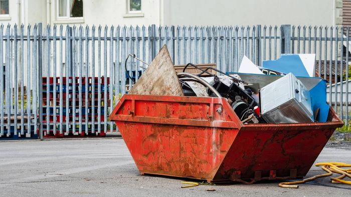 A red dumpster filled with trash is parked in front of a fence.