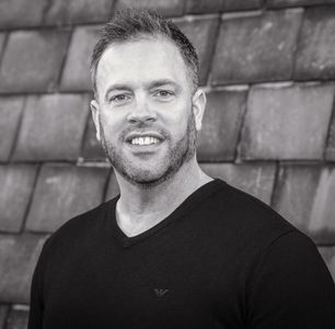 A man with a beard is wearing a black shirt and smiling in a black and white photo.