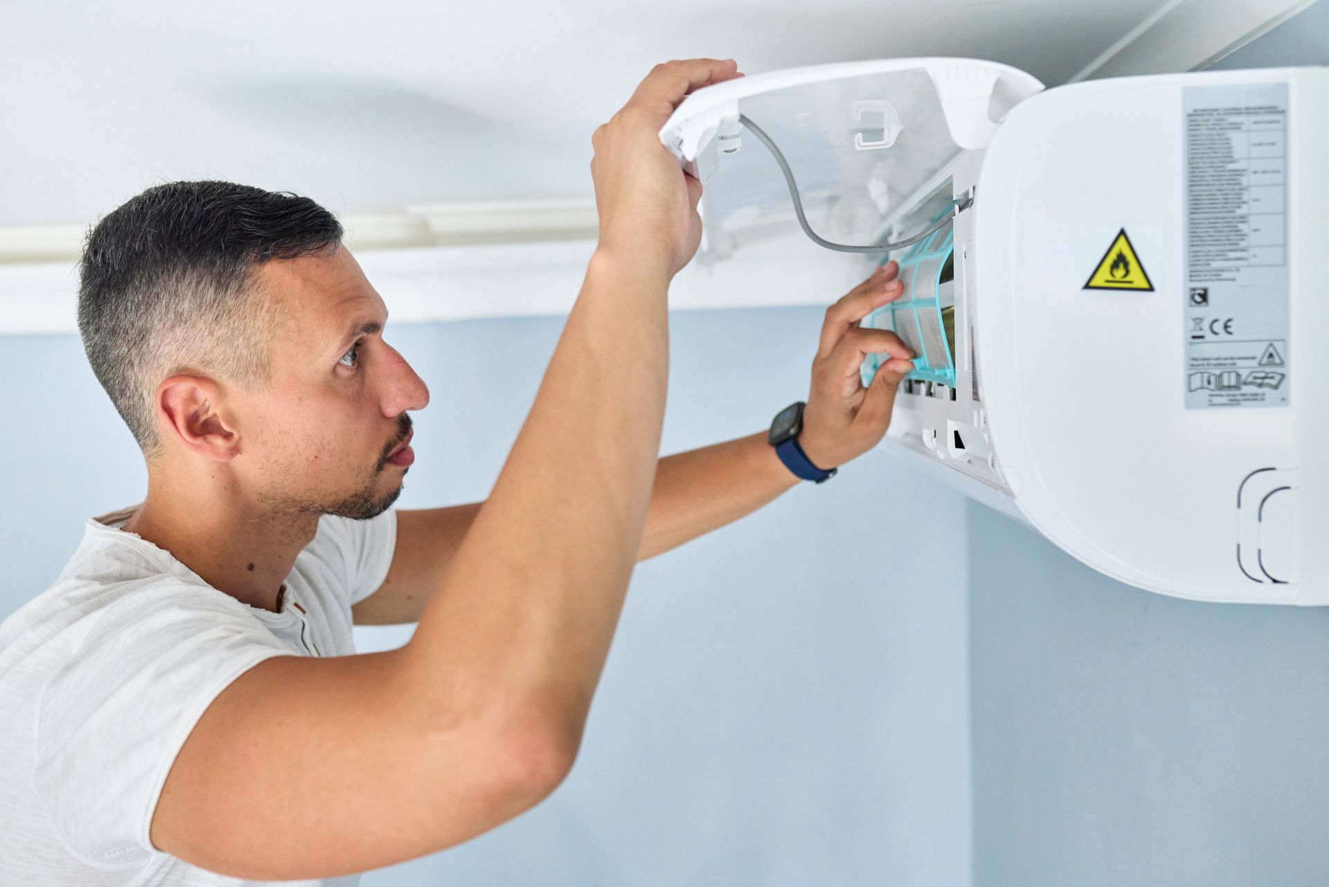 A man, wearing a white tee, services an air conditioning unit inside a room.
