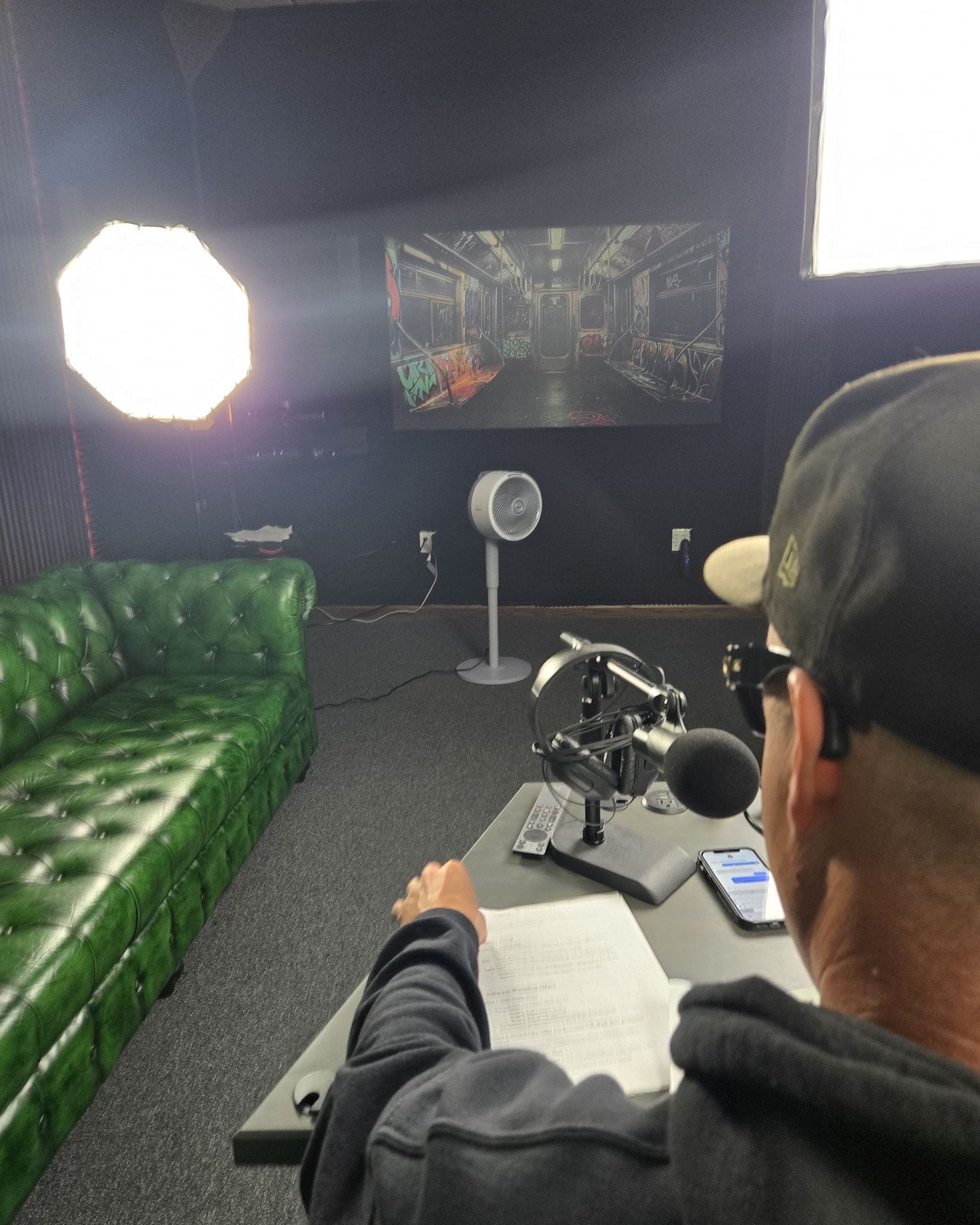 Person at a desk in a studio, facing a camera and microphone with a green couch and bright light in the background