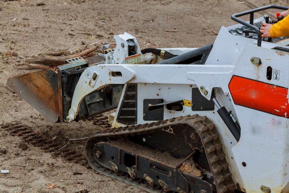 White skid steer with tracked wheels scoops dirt on a muddy construction site — Old Mates Grass Cutters in Deeragun, QLD