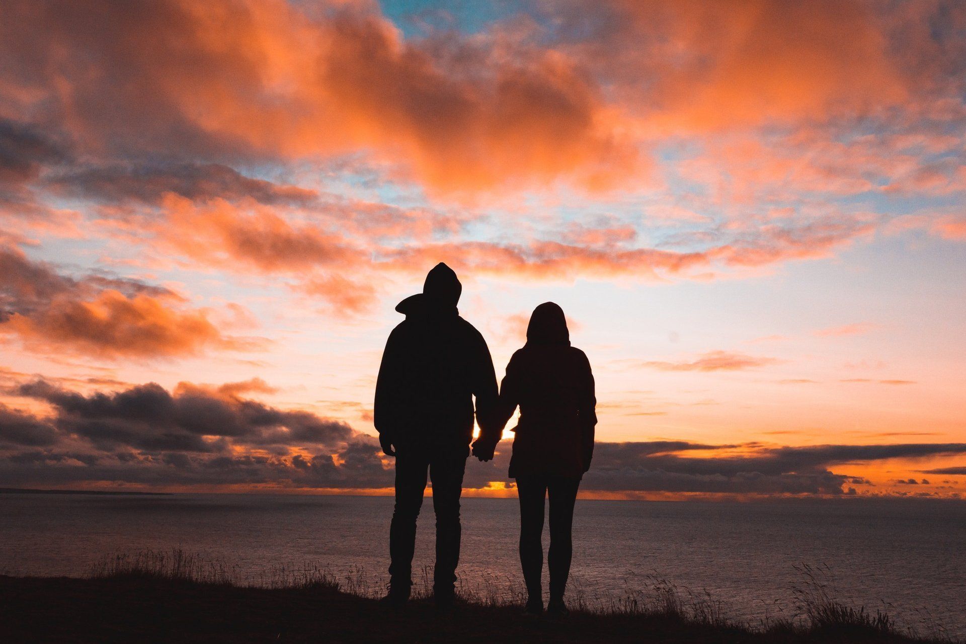 A man and a woman are holding hands while standing on top of a hill overlooking the ocean at sunset.