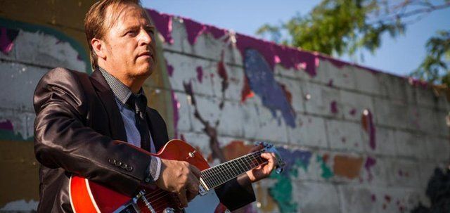 A man in a suit and tie is playing a guitar in front of a graffiti wall.
