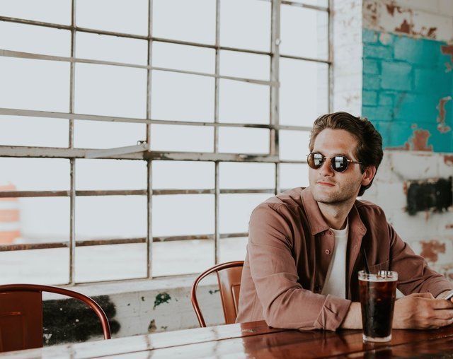A man wearing sunglasses is sitting at a table with a glass of soda.
