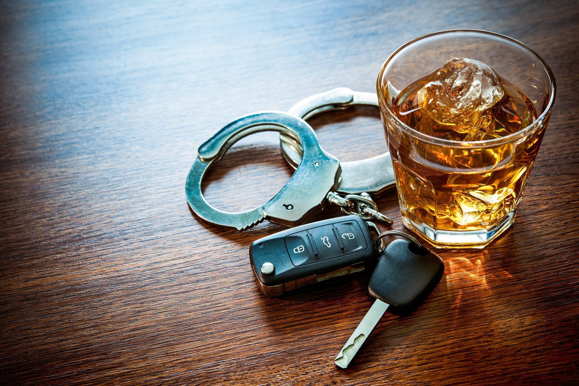 A pair of handcuffs , a car key and a glass of whiskey on a wooden table.