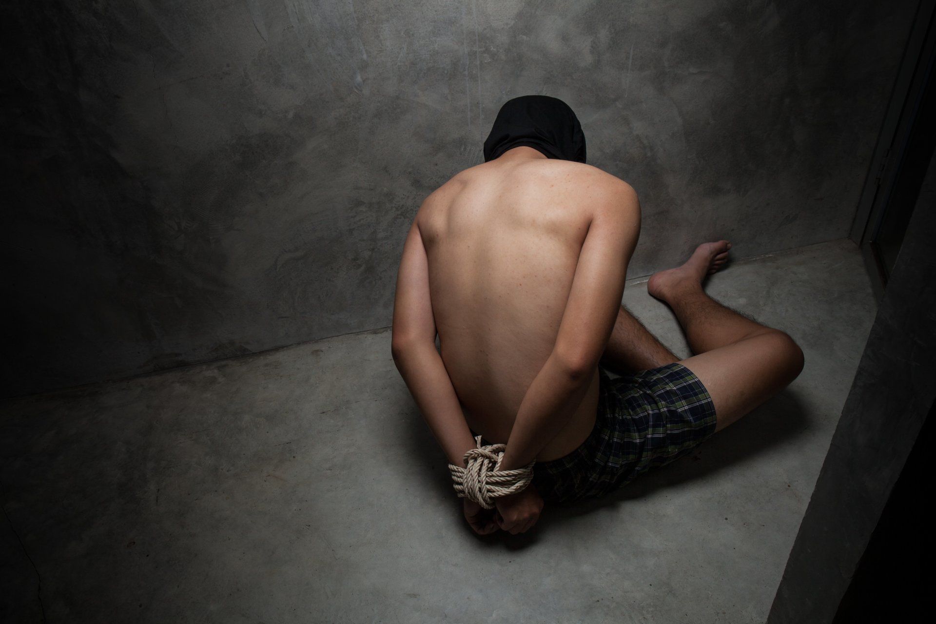 A shirtless man is tied up and sitting on the floor in a dark room.