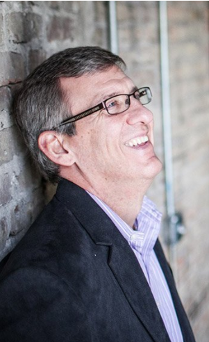 A man wearing glasses and a suit is leaning against a brick wall and smiling.