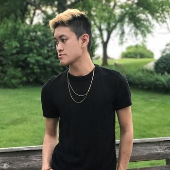 A young man wearing a black shirt and a gold chain is standing next to a wooden fence.