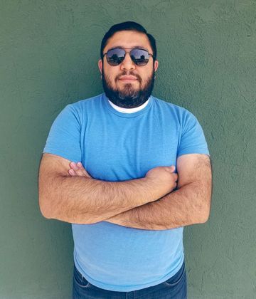 a man with a beard wearing sunglasses and a blue shirt is standing with his arms crossed .