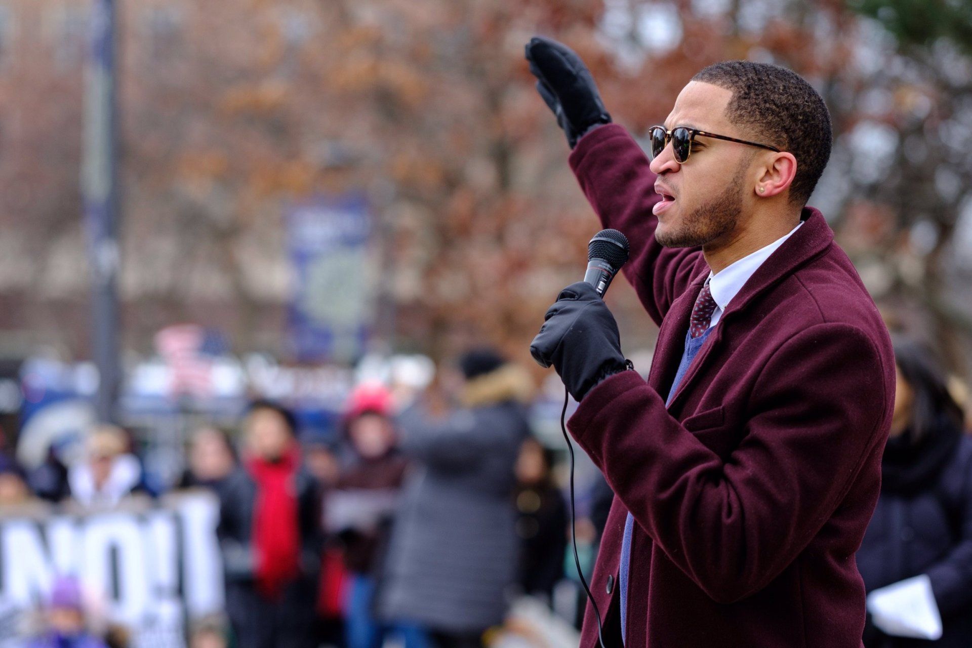 A man in a maroon coat is holding a microphone and giving a speech.