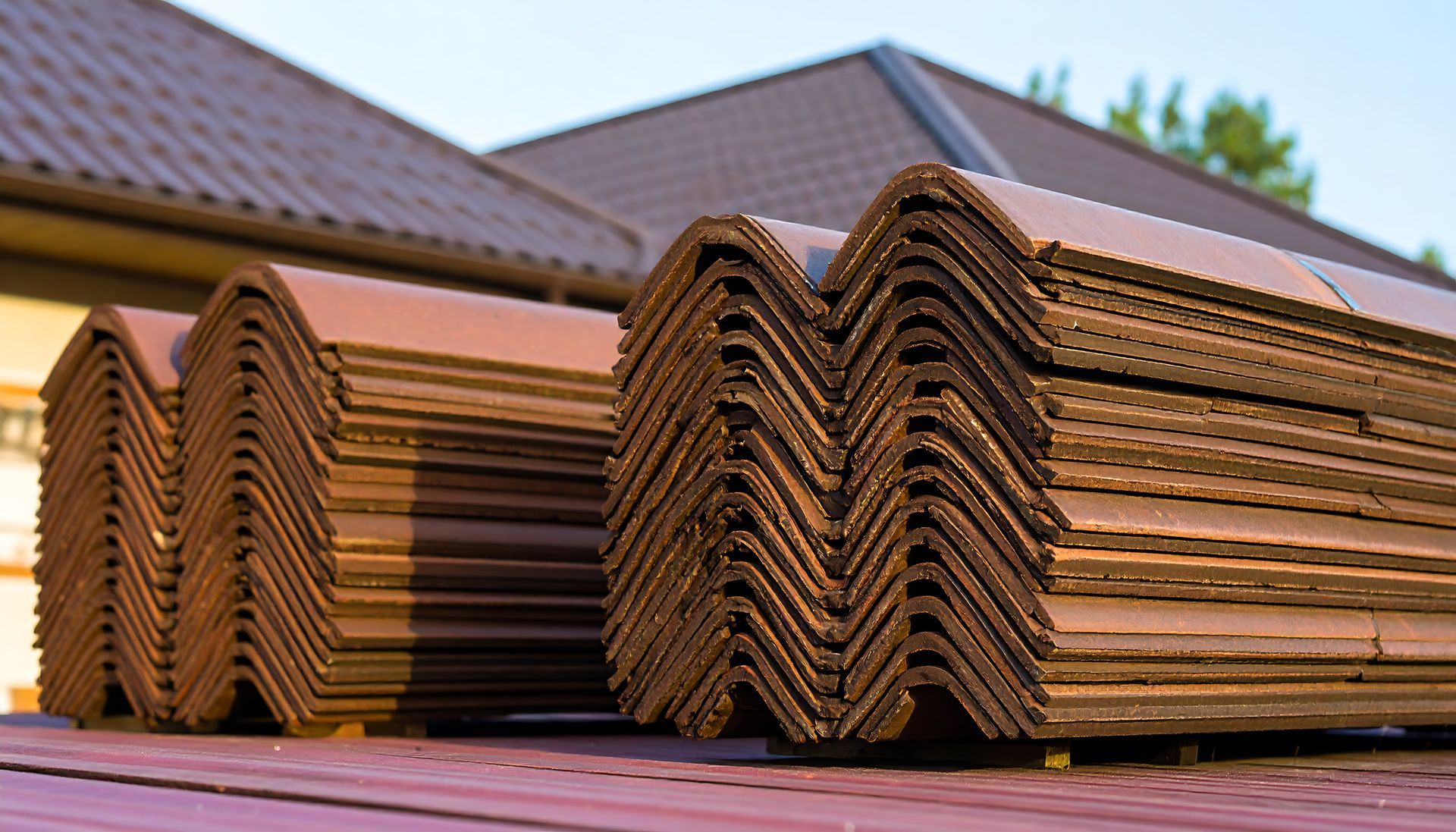 Stacked brown roofing tiles sit on a rooftop surface against a backdrop of a house