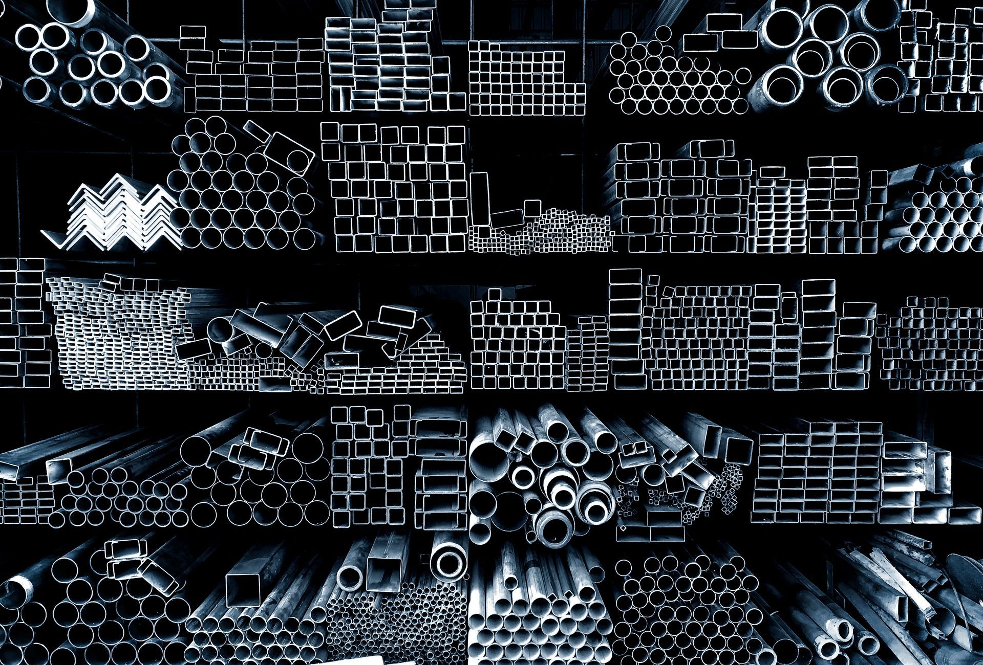 Assorted steel pipes, beams, and metal profiles neatly stacked in a warehouse or industrial storage area.