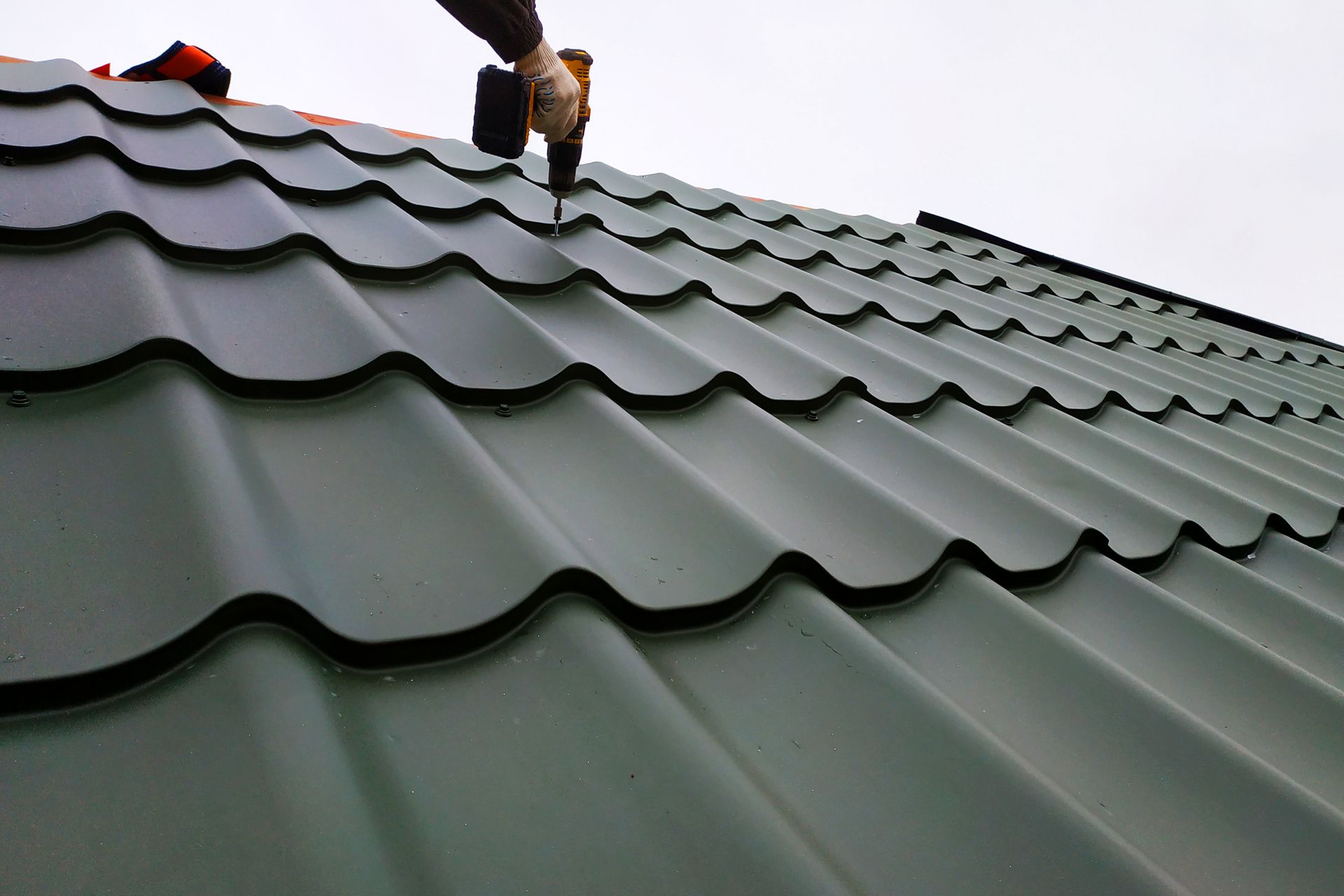 Close-up of metal roofing on a building, with a hand drilling into one of the metal tiles.