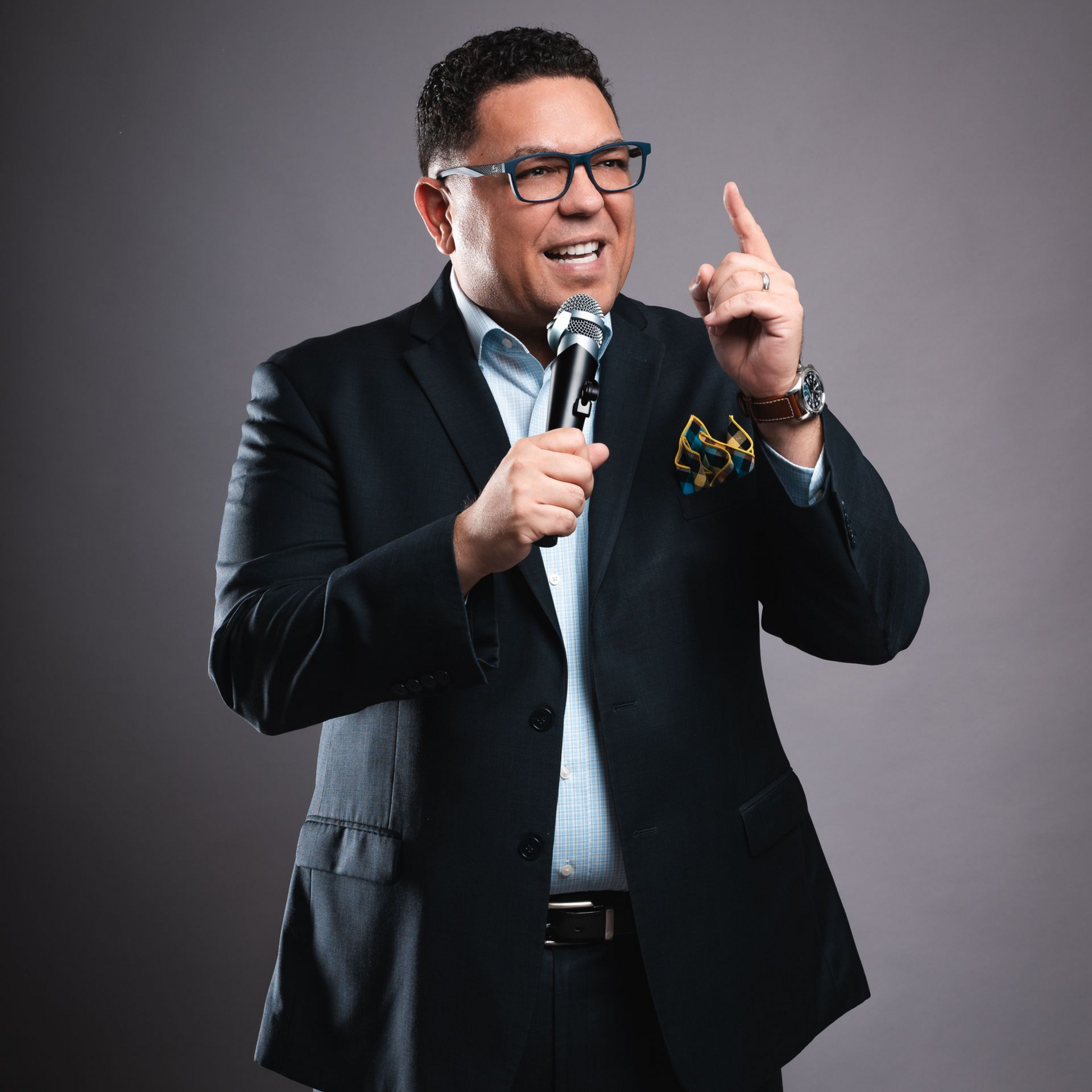 Dr. Peter Rios holding a microphone, pointing upward. He wears glasses, smiling in a studio setting.