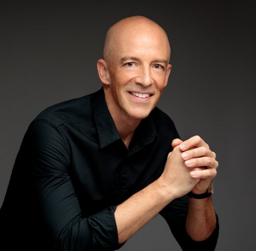 Michael Ceely, high-performance executive coach, smiling, wearing a black shirt, resting hands together on a dark background.